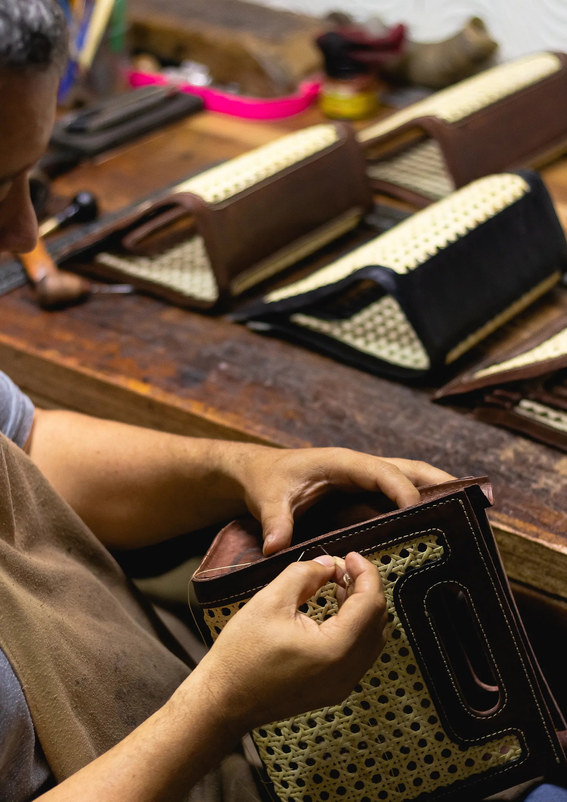 Una mujer tejiendo bolsas de hojas de palma de iraca marrón natural con un par de estas bolsas terminadas detrás de ella
